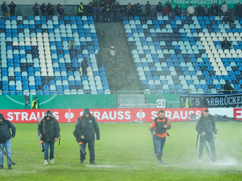 Im Ludwigspark-Stadion versuchen Helfer, Wasserlachen vom Spielfeld zu entfernen. - Foto: Uwe Anspach/dpa