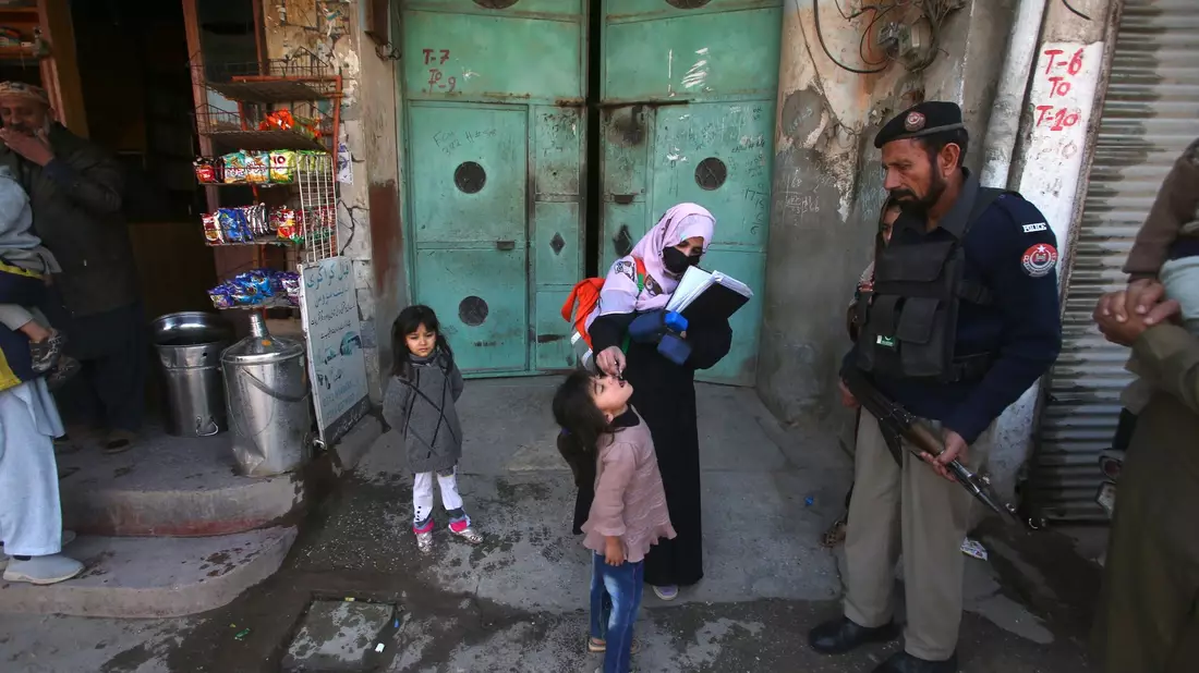 Ein Polizist in Pakistan steht Wache, während einem Kind ein Polio-Impfstoff verabreicht wird. - Bild: Muhammad Sajjad/AP/dpa