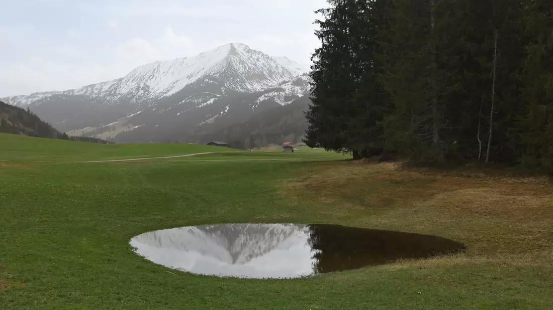Die im Dunst liegenden Alpen spiegeln sich in einem Weiher. Der Saharastaub-Höhepunkt ist überschritten. - Bild: Karl-Josef Hildenbrand/dpa