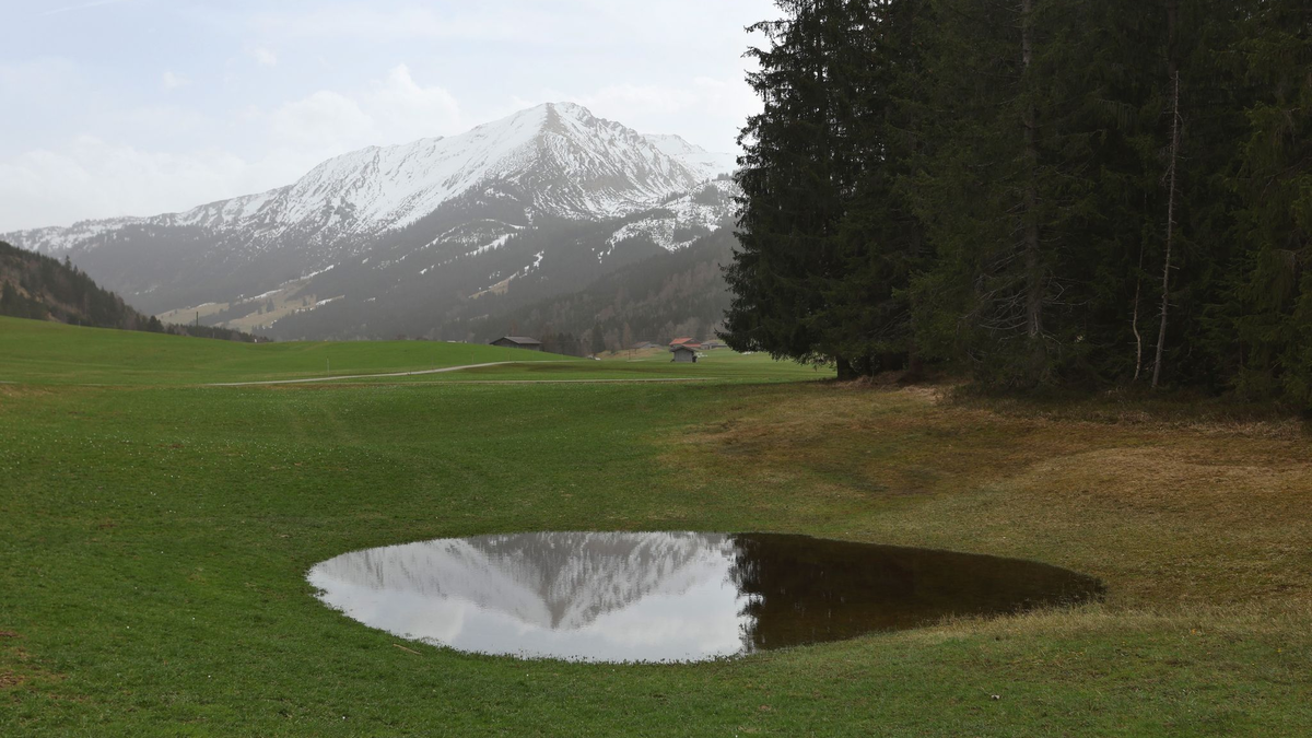 Die im Dunst liegenden Alpen spiegeln sich in einem Weiher. Der Saharastaub-Höhepunkt ist überschritten. - Foto: Karl-Josef Hildenbrand/dpa