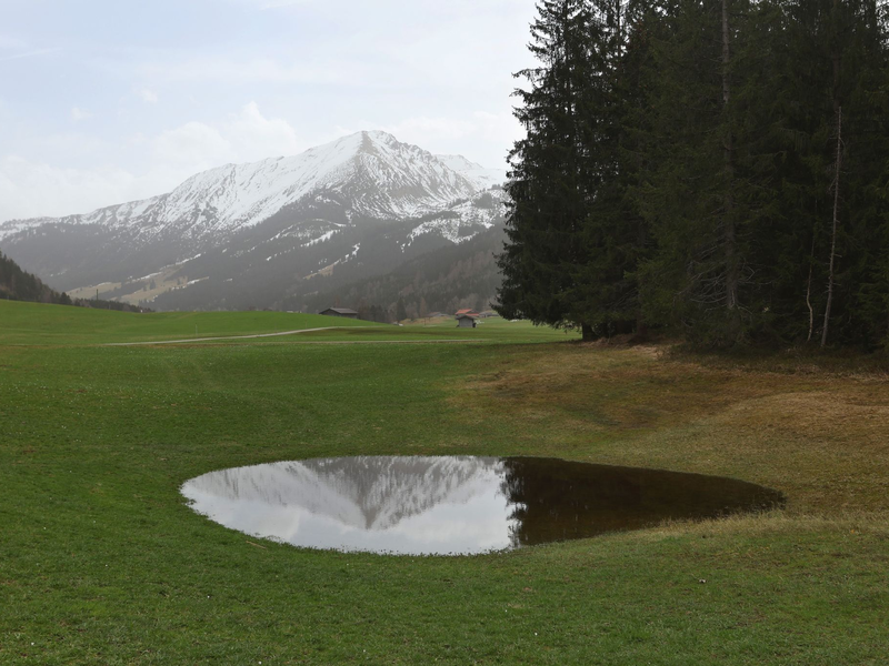 Die im Dunst liegenden Alpen spiegeln sich in einem Weiher. Der Saharastaub-Höhepunkt ist überschritten. - Foto: Karl-Josef Hildenbrand/dpa
