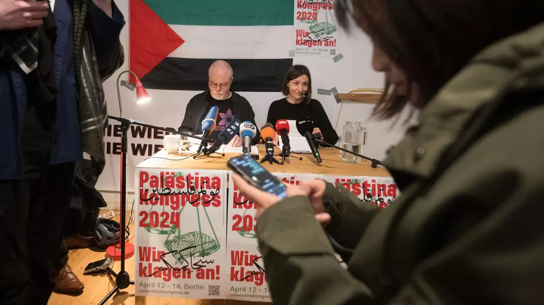 Teilnehmer sitzen vor Beginn des Palästina-Kongress 2024 im Veranstaltungsraum vor einem Plakat des Kongresses. - Bild: Sebastian Christoph Gollnow/dpa