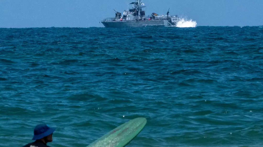 Ein Surfer wartet auf eine Welle, während ein israelisches Marineschiff im Mittelmeer vor der Küste von Hadera patrouilliert. - Bild: Ariel Schalit/AP