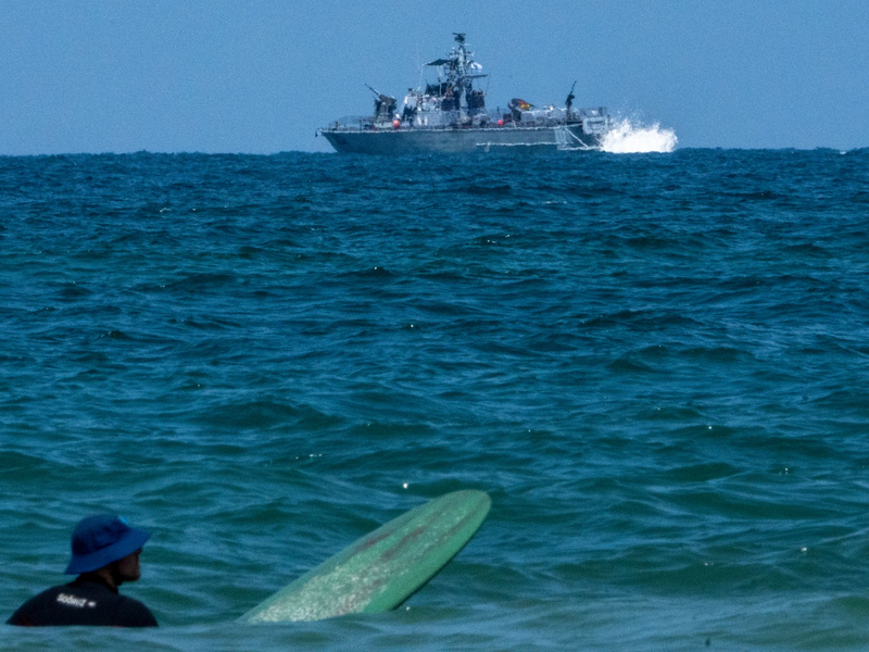 Ein Surfer wartet auf eine Welle, während ein israelisches Marineschiff im Mittelmeer vor der Küste von Hadera patrouilliert. - Foto: Ariel Schalit/AP