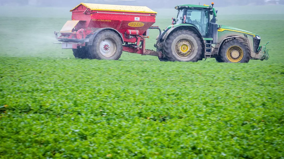 Ein Traktor fährt mit einem Streuwagen über ein Feld und bringt Dünger aus. - Bild: dpa