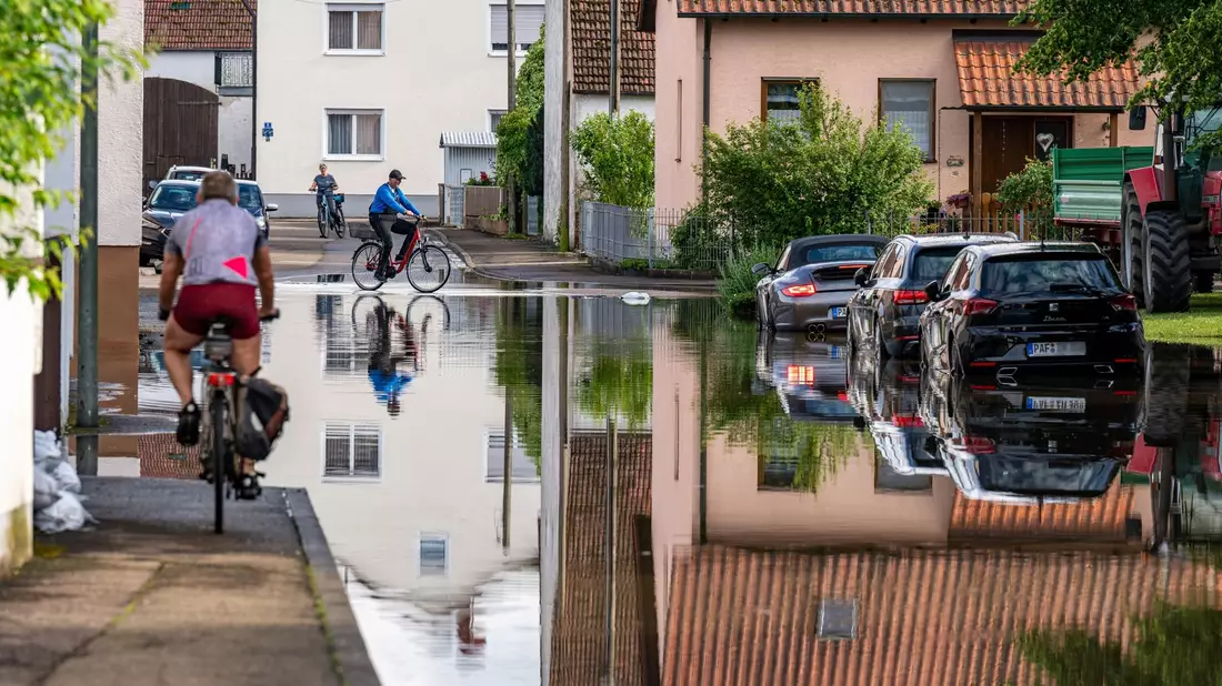 Radler fahren durch eine überflutete Straße. Im Bereich der Gemeinde Baar-Ebenhausen war ein Damm an zwei Stellen geborsten. - Bild: Armin Weigel/dpa