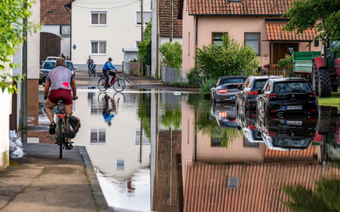 Radler fahren durch eine überflutete Straße. Im Bereich der Gemeinde Baar-Ebenhausen war ein Damm an zwei Stellen geborsten. - Foto: Armin Weigel/dpa