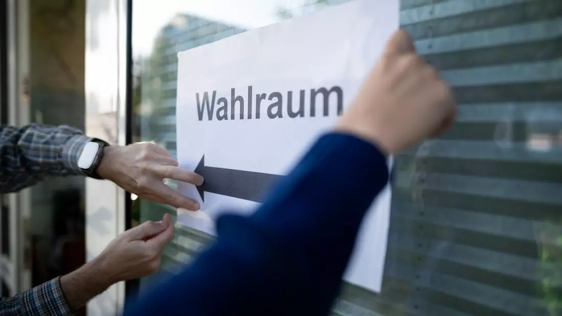 Wahlhelfer hängen einen Zettel, auf dem „Wahlraum“ steht, auf. - Bild: Fabian Strauch/dpa