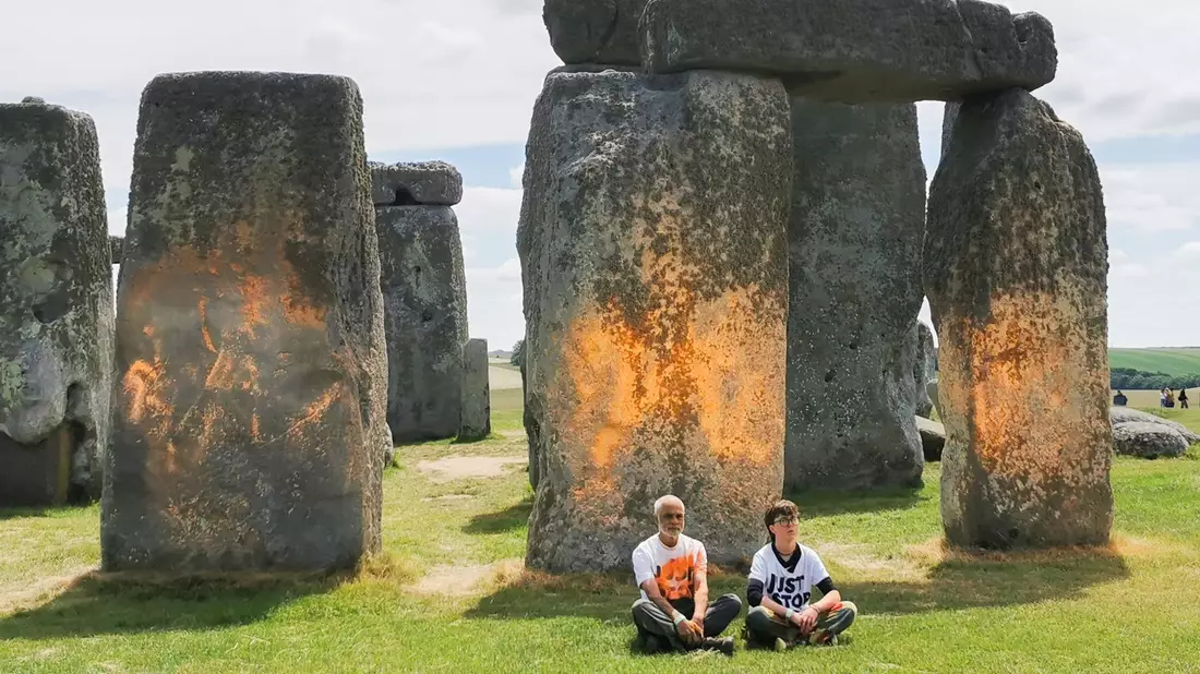 Demonstranten von «Just Stop Oil» sitzen vor dem Steinmonument Stonehenge, das sie zuvor mit orangener Farbe besprüht haben. - Bild: Just Stop Oil/PA Media/dpa