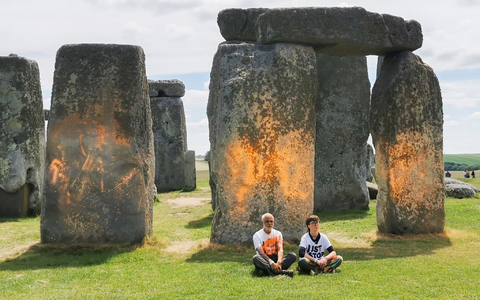 Demonstranten von «Just Stop Oil» sitzen vor dem Steinmonument Stonehenge, das sie zuvor mit orangener Farbe besprüht haben. - Foto: Just Stop Oil/PA Media/dpa