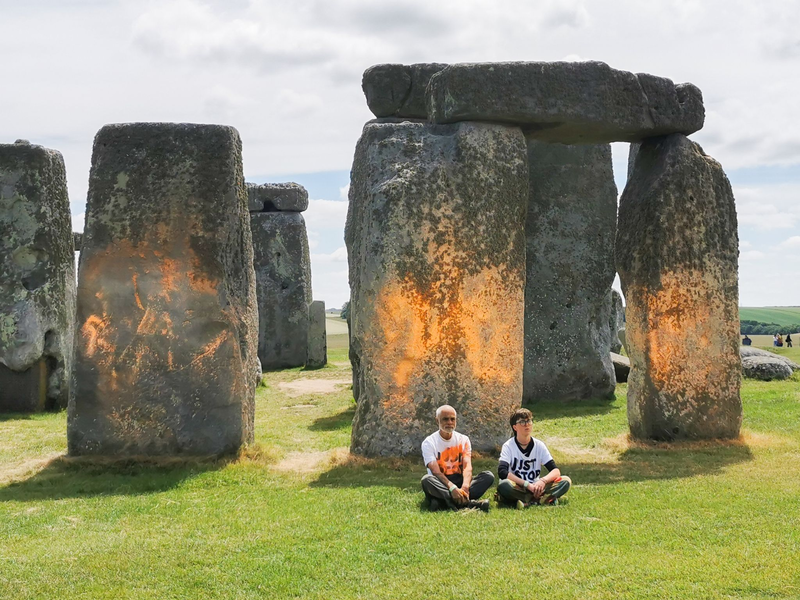 Demonstranten von «Just Stop Oil» sitzen vor dem Steinmonument Stonehenge, das sie zuvor mit orangener Farbe besprüht haben. - Foto: Just Stop Oil/PA Media/dpa