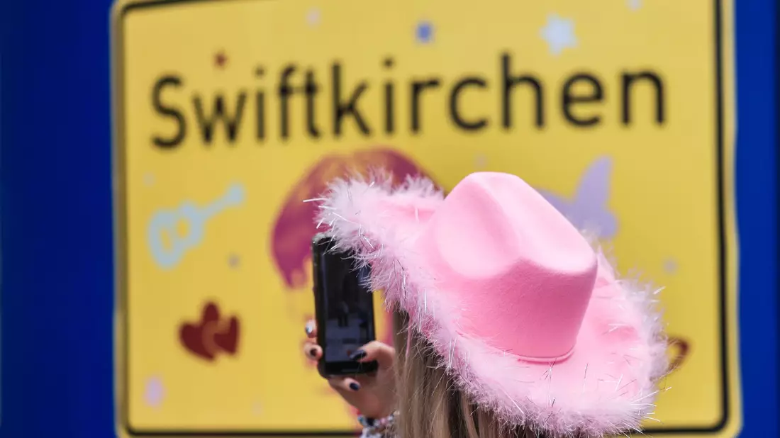 Ein Fan fotografiert vor dem Konzert der Sängerin Taylor Swift in Gelsenkirchen ein «Swiftkirchen»-Schild. Während des Konzerts am Abend zog der Datenverbrauch in den Handynetzen in der Veltins Arena kräftig an. - Bild: Oliver Berg/dpa