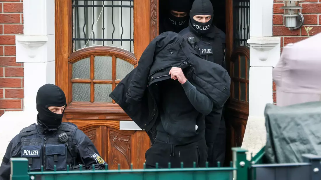 Polizeibeamte führen einen Tatverdächtigen aus einem durchsuchten Haus.  - Bild: Christoph Reichwein/dpa