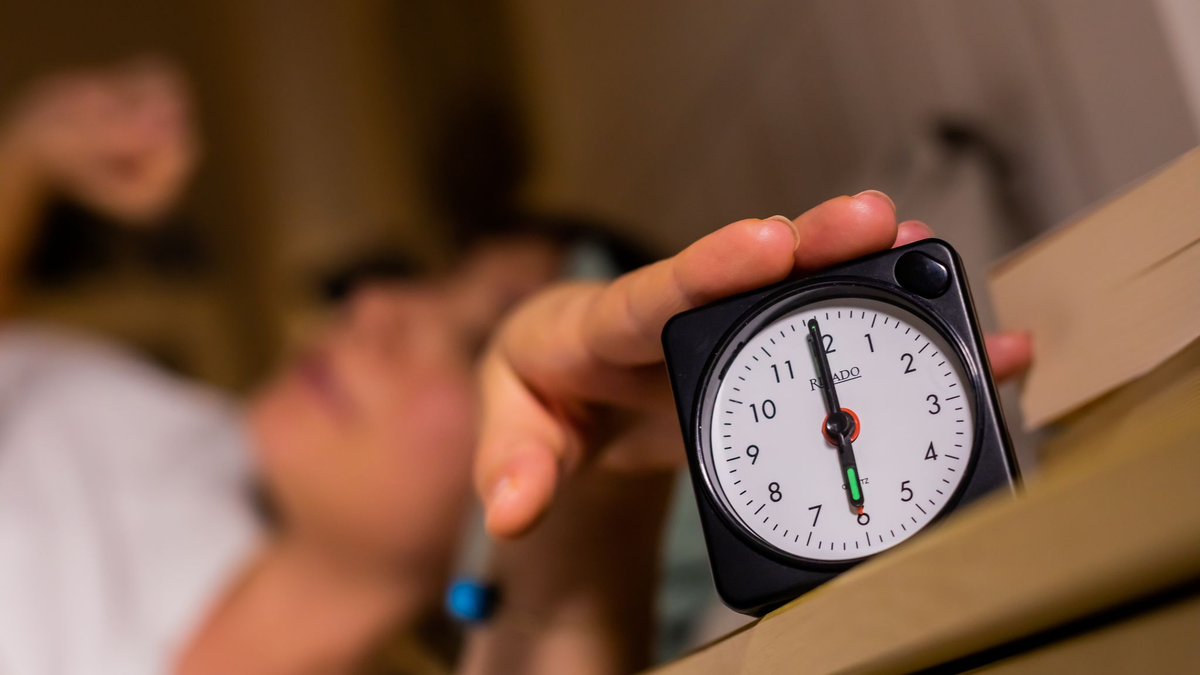 Eine Frau schaltet vom Bett aus einen auf sechs Uhr gestellten Wecker aus.  - Foto: Christoph Soeder/dpa