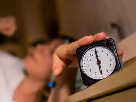 Eine Frau schaltet vom Bett aus einen auf sechs Uhr gestellten Wecker aus.  - Foto: Christoph Soeder/dpa