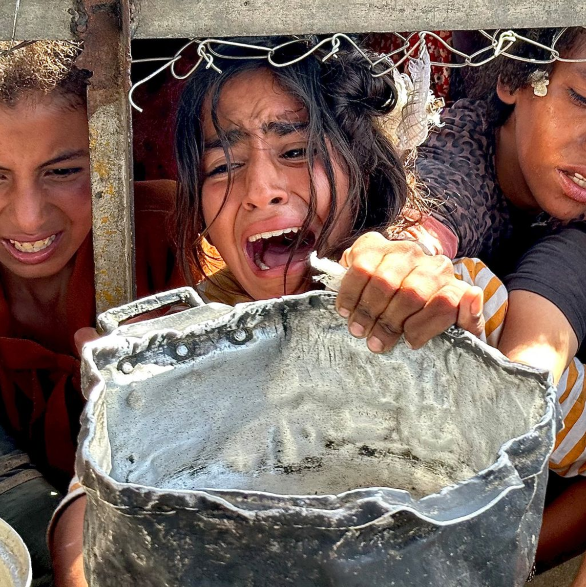 Palästinensische Kinder versuchen im Gebiet Al-Mawasi im Gazastreifen, mit ihren Töpfen Nahrungsmittel zu bekommen, die von Wohltätigkeitsorganisationen verteilt werden. - Foto: Moaz Abu Taha/APA Images via ZUMA Press Wire/dpa