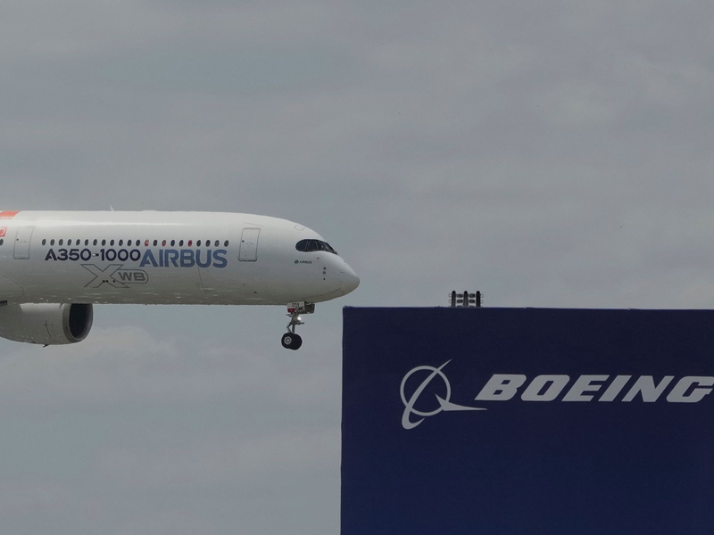 Die zwei großen Konkurrenten: Ein Airbus fliegt bei einer Vorführung auf der Luftfahrtmesse Salon du Bourget an einem Logo von Boeing vorbei. - Foto: Michel Euler/AP/dpa