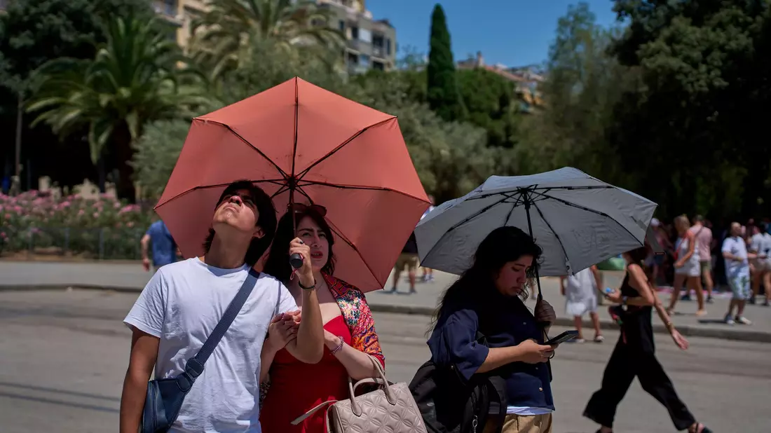 Touristen schützen sich amvor der Kirche Sagrada Familia mit Regenschirmen vor der Sonne.  - Bild: Emilio Morenatti/AP/dpa