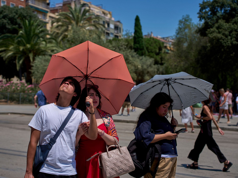 Touristen schützen sich amvor der Kirche Sagrada Familia mit Regenschirmen vor der Sonne.  - Foto: Emilio Morenatti/AP/dpa