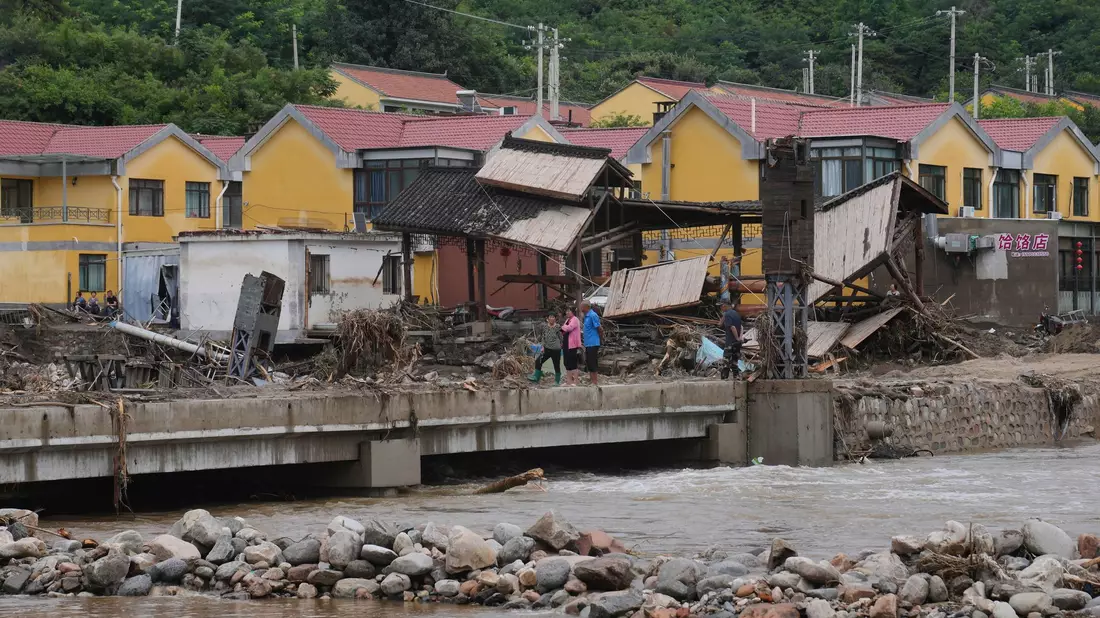 Dorfbewohner versammeln sich in der Nähe einer Brücke, die nach schweren Regenfällen im Bezirk Miyun am Stadtrand von Peking überflutet wurde. - Bild: Andy Wong/AP/dpa