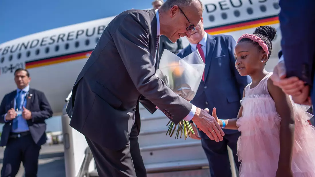 Kanzler Friedrich Merz wird auf dem Flughafen von Johannesburg von einem Mädchen begrüßt.  - Bild: Michael Kappeler/dpa