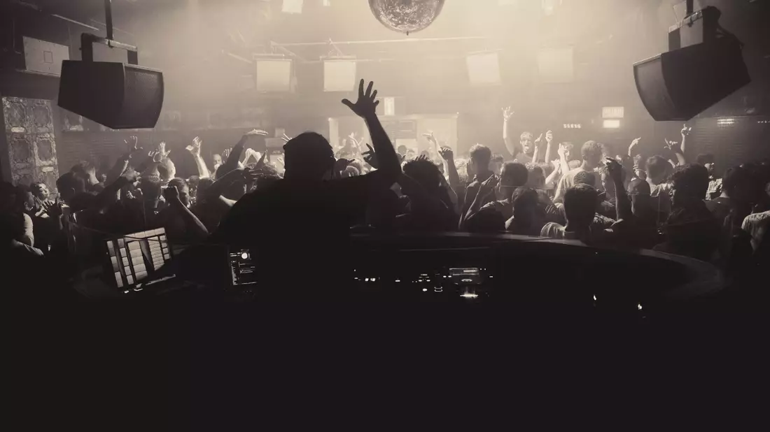 DJ am Pult vor tanzender Menge unter Diskokugel im neblig-dunstigen Club
