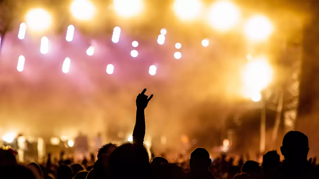 Hand mit Rockgeste vor verschwommener Bühne in warmem Gelb-Pink beim Festival