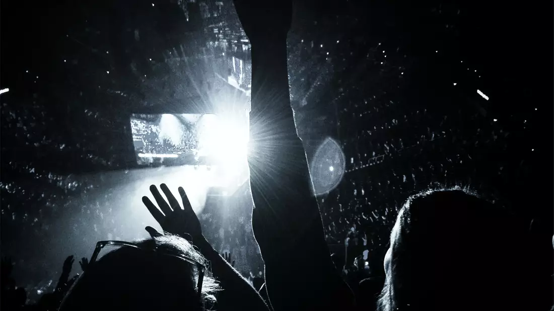 Erhobene Hände vor heller Bühne mit Videoleinwand in voller Arena in Schwarzweiß