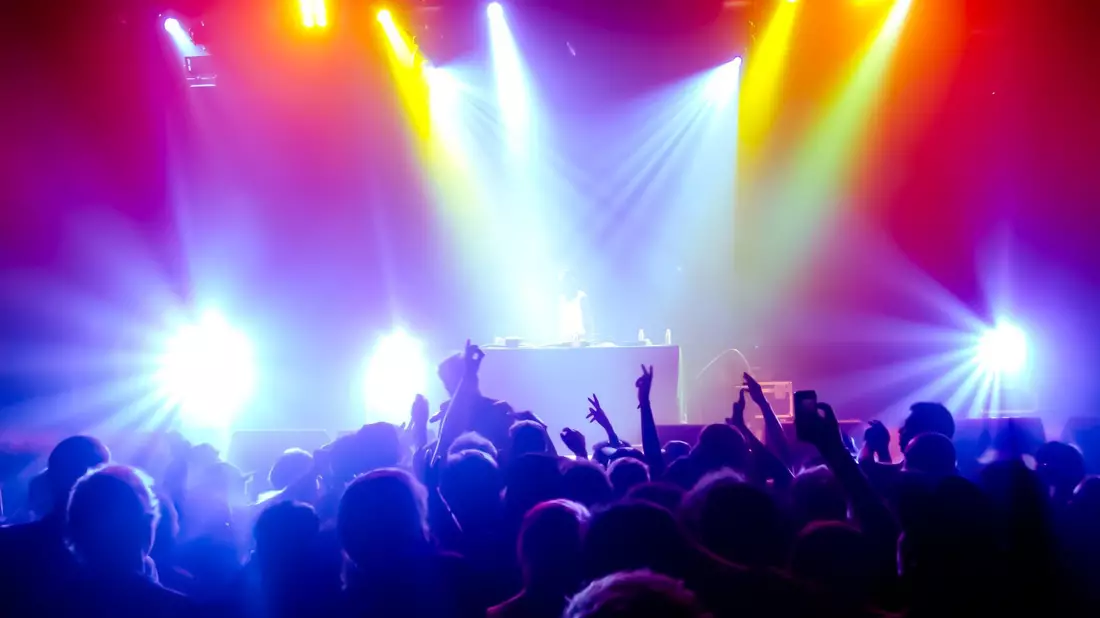 DJ auf Bühne vor tanzender Menge mit bunter Lichtshow in Rot, Gelb und Violett
