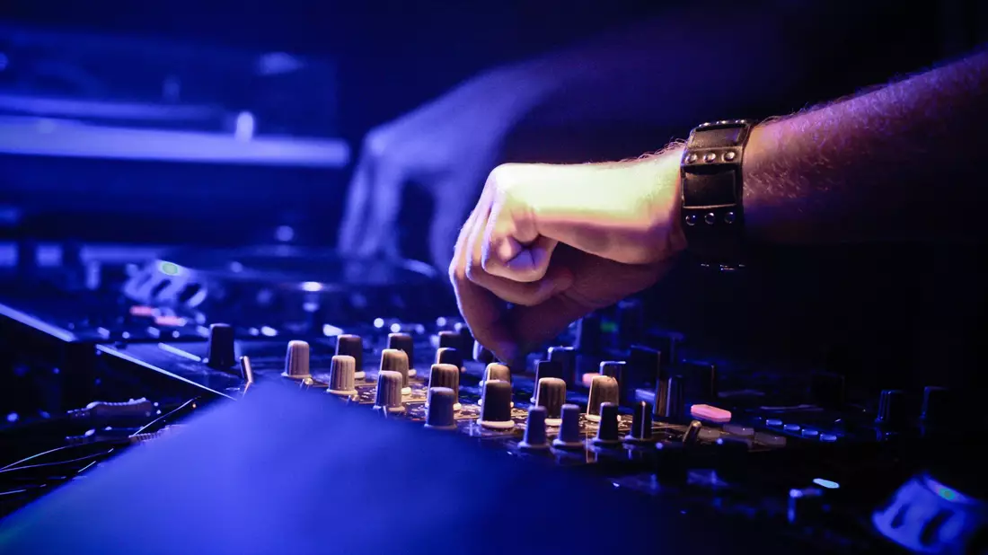 Hände eines DJs bedienen Regler und Fader am Mischpult im blauen Clublicht