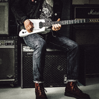 Rockige Pose im Backstage-Look: Auf einem Verstärker sitzend spielt der Gitarrist seine weiße E-Gitarre, umringt von Amps. - Foto: THN