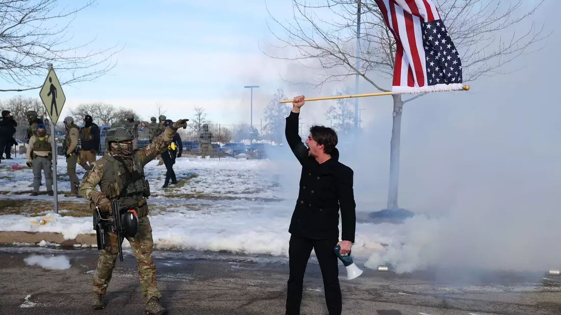 Ein Protestierender mit einer amerikanischen Flagge steht einem ICE-Beamten gegenüber. - Bild: Adam Bettcher/AP/dpa