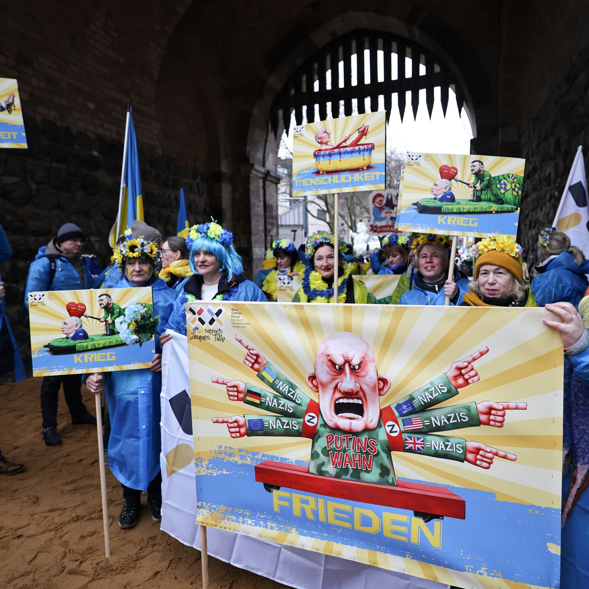 Der ukrainische Hilfsverein Blau-Gelbes-Kreuz hält Plakate mit Putin-Motiven hoch. Das ist als Zeichen der Solidarität mit Wagenbauer Jacques Tilly gemeint. Dieser wird in Russland strafrechtlich verfolgt.  - Foto: Oliver Berg/dpa