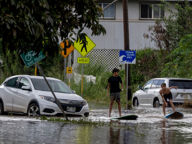 Zwei Jugendliche surfen in Waialua neben einem liegengebliebenen Fahrzeug im Hochwasser. - Foto: Stephen Lam/San Francisco Chronicle via AP/dpa