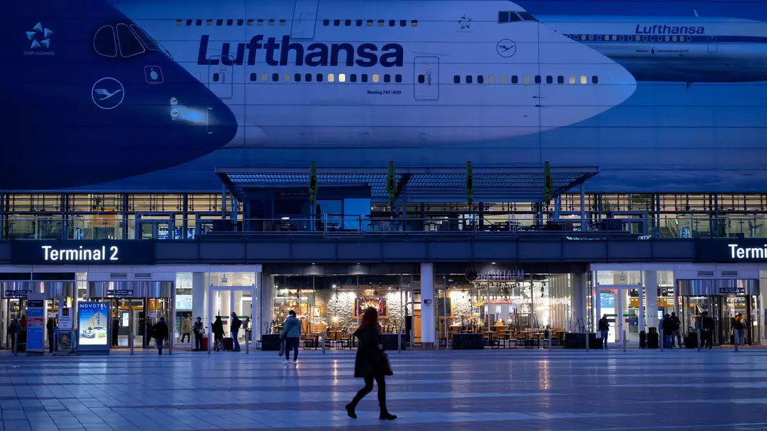Passagiere gehen am Münchner Flughafen vor dem Terminal 2 an einem Plakat mit Flugzeugen der Lufthansa entlang. - Bild: Sven Hoppe/dpa