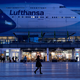 Passagiere gehen am Münchner Flughafen vor dem Terminal 2 an einem Plakat mit Flugzeugen der Lufthansa entlang. - Foto: Sven Hoppe/dpa