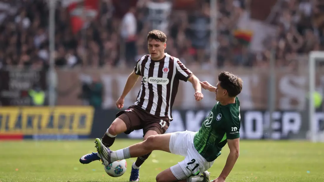 Abstiegskampf pur: Der Wolfsburger Joakim Maehle packt die Grätsche gegen St. Paulis Arkadiusz Pyrka aus. - Bild: Christian Charisius/dpa