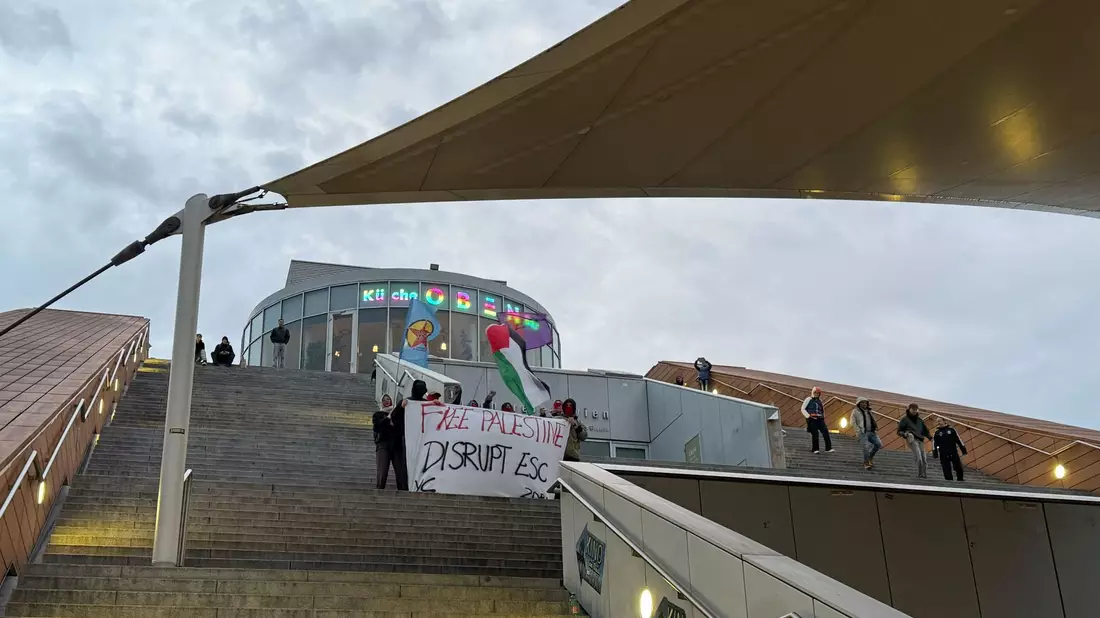 Die Aktivisten protestierten mit Sprechchören und Plakaten auf den Stufen der städtischen Hauptbücherei gegen die Teilnahme Israels am ESC.  - Bild: Britta Schultejans/dpa