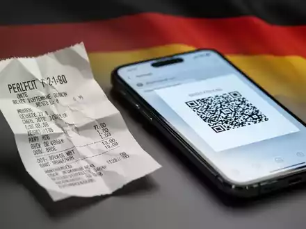 Crumpled paper receipt next to a smartphone showing a digital receipt, with a blurred German flag in the background. - Foto: über boerse-global.de