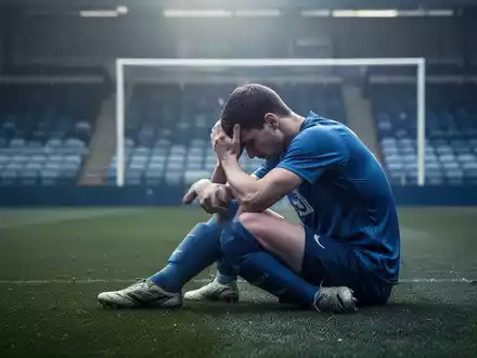 Ein Fußballspieler sitzt allein auf einem leeren Spielfeld, den Kopf in den Händen, was Nachdenklichkeit und Verletzlichkeit symbolisiert. - Foto: über boerse-global.de