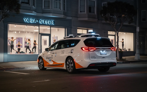 A white Waymo robotaxi with orange accents drives away from a modern yoga studio at night in San Francisco. - Foto: über boerse-global.de