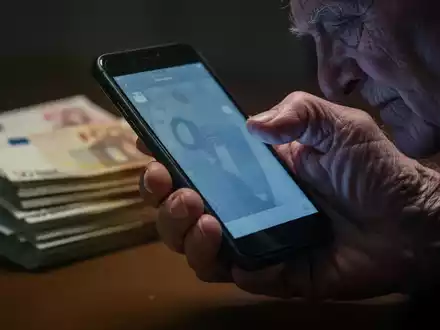 Nahaufnahme einer älteren Hand, die ein Smartphone hält, mit einem Stapel Euro-Banknoten im unscharfen Hintergrund. - Foto: über boerse-global.de
