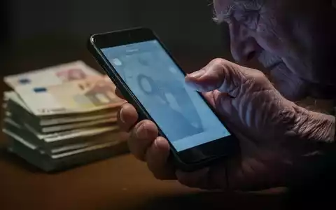 Nahaufnahme einer älteren Hand, die ein Smartphone hält, mit einem Stapel Euro-Banknoten im unscharfen Hintergrund. - Foto: über boerse-global.de