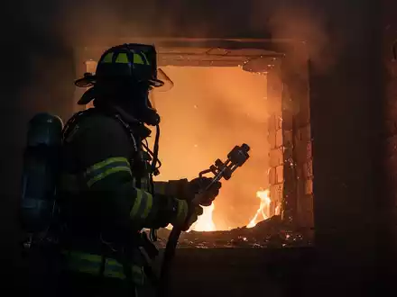 Ein Feuerwehrmann in voller Ausrüstung vor einem rauchenden Schornstein, der ein Werkzeug hält. Orangefarbenes Feuer leuchtet im Schornstein. - Foto: über boerse-global.de