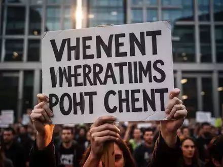 Nahaufnahme eines Protestschildes, das von einem Demonstranten gehalten wird, mit einem unscharfen Hintergrund eines Unternehmensgebäudes. - Foto: über boerse-global.de