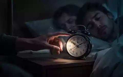 Eine Hand greift nach einem wecker auf einem Nachttisch in einem dunklen Schlafzimmer, der die frühen Morgenstunden anzeigt. - Foto: über boerse-global.de