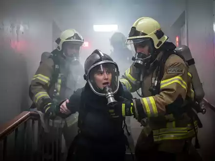 Ein Feuerwehrmann führt eine Person mit einer Rauchschutzhaube durch ein verrauchtes Treppenhaus. Rauch und Notbeleuchtung dominieren. - Foto: über boerse-global.de