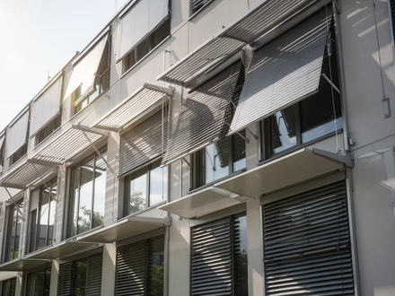 Eine moderne Gebäudefassade mit ausgefahrenen externen Sonnenschutzsystemen und großen Fenstern, die auf passive Kühlung hinweisen. - Foto: über boerse-global.de