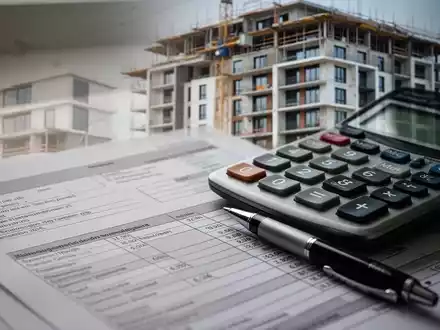 Nahaufnahme von Finanzdokumenten und einem Taschenrechner, im Hintergrund unscharf ein modernes Wohngebäude im Bau. - Foto: über boerse-global.de