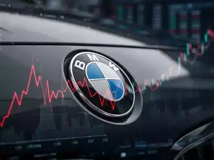 Nahaufnahme eines BMW-Logos auf einem dunklen Auto, das eine rote, fallende Linie widerspiegelt, im Hintergrund unscharfe Finanzdaten. - Foto: über boerse-global.de